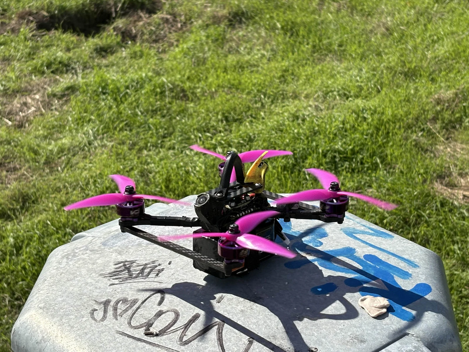 FPV quadcopter with magenta propellers and a yellow camera mount outdoors