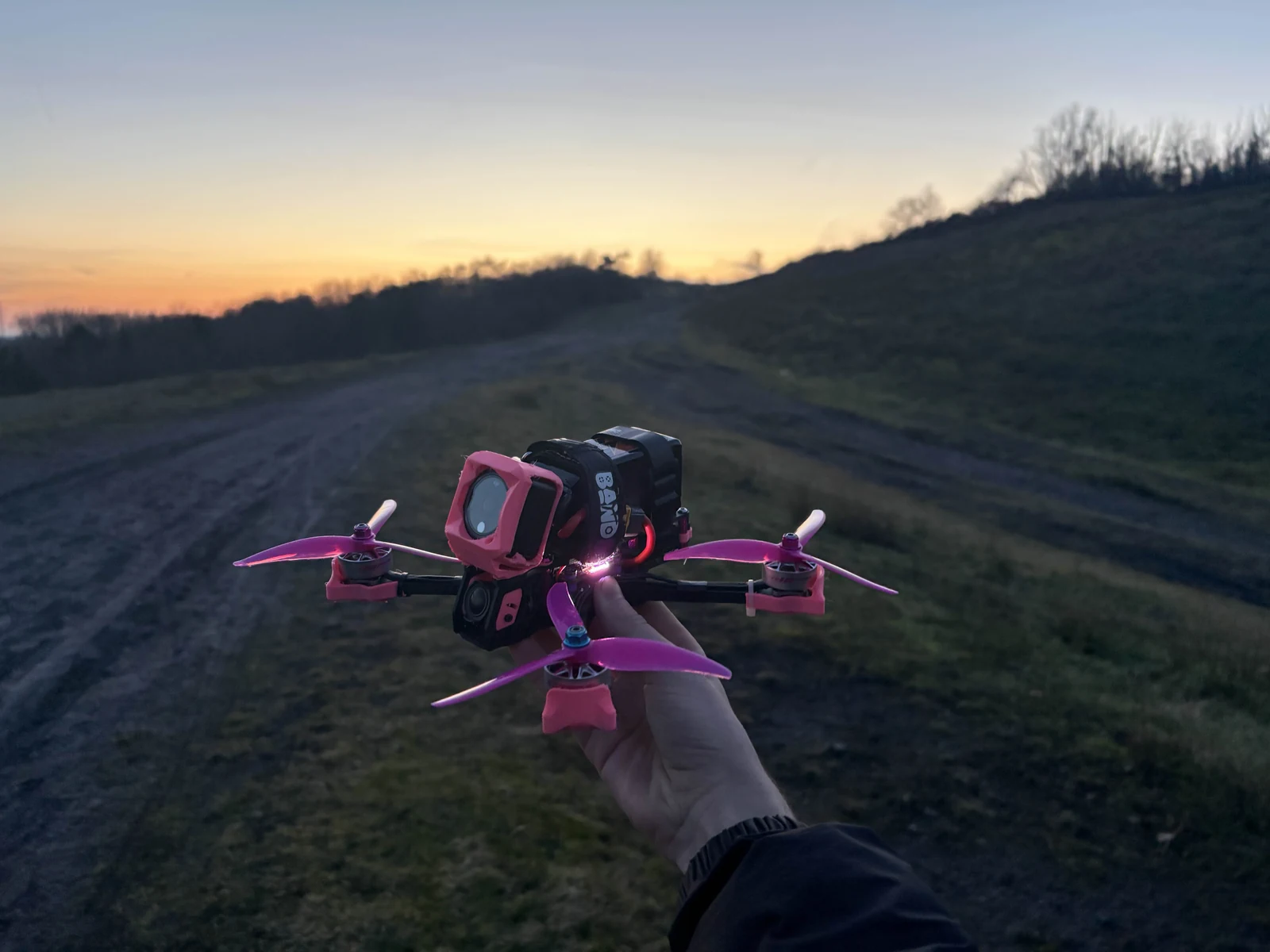 Hand-held pink-accented FPV drone against a sunset landscape